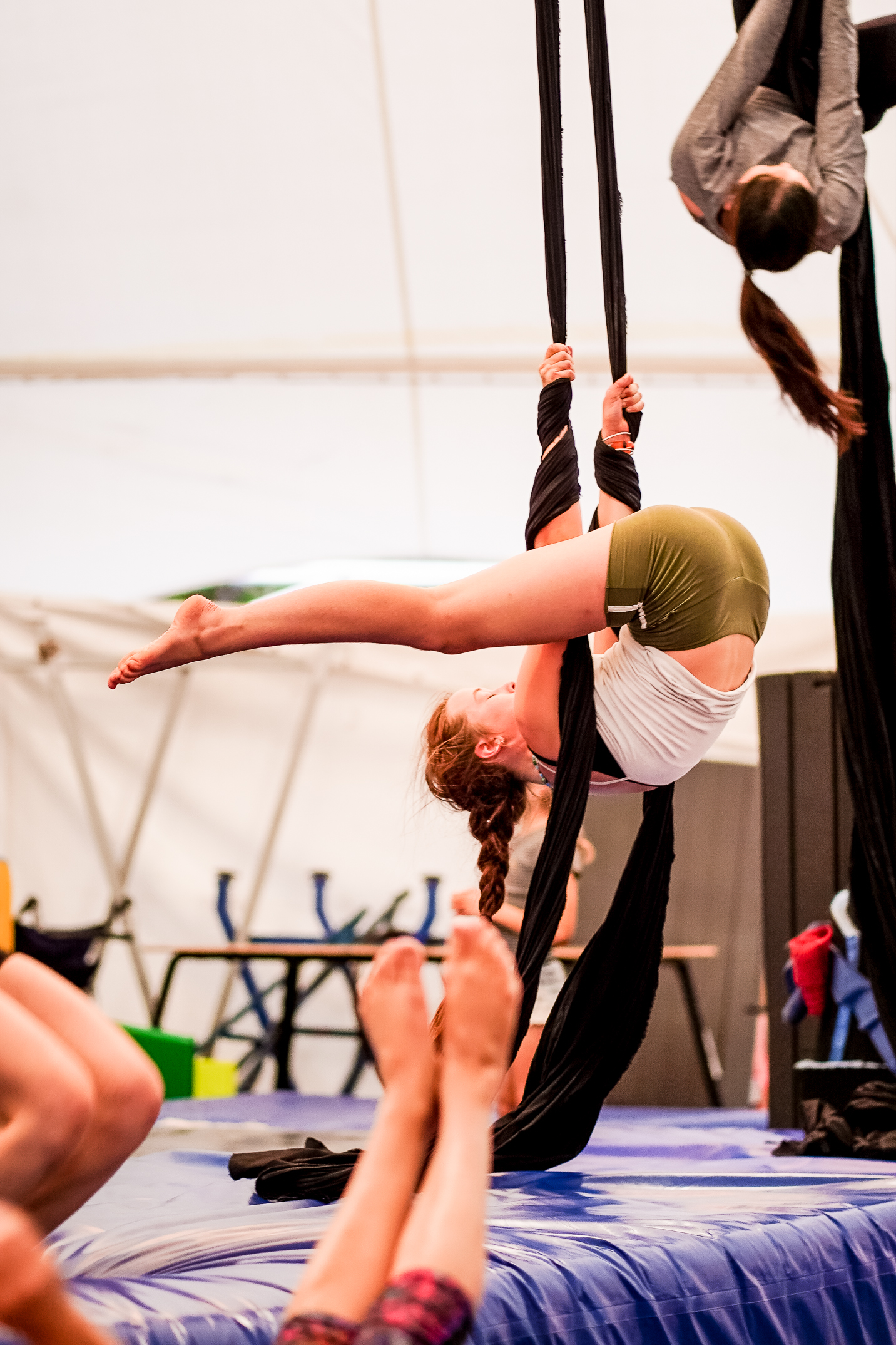 one person is inverted on an aerial silk low to the ground, people stretching can be seen in the background